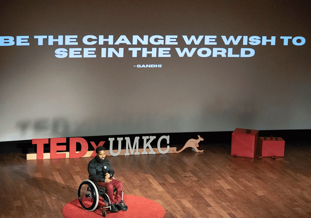 Wes wearing maroon pants, black shoes, and shiny black shirt, on a large stage with TED Talks logo and UMKC logo behind him and the words 